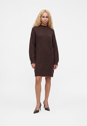 Femme aux longs cheveux blonds bouclés portant une robe-pull tricotée marron foncé et des chaussures à talons noirs, debout sur un fond blanc.