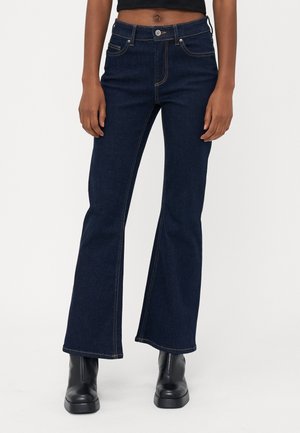 Osoba ubrana w ciemnoniebieskie dzwony z wysokim stanem, czarne botki za kostkę oraz czarny krótki top, stojąca na tle białego tła.