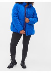 Blå puffjakke med høj krave, quiltet tekstur, lynlås fortil, båret over en lilla top, parret med sorte stramme jeans og sorte combat støvler.
