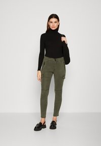 Oljčnozelene cargo hlače z zadrgo na sprednjih žepih, v kombinaciji s črnimi rebrastimi turtleneck majicami in črnimi platformami loafers.