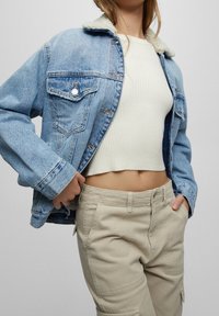 Denim-takk med en lys blå nyanse, skjærkollor, knappelukking foran, og to brystlommer, sammen med en hvit ribbet crop-top og beige bukser.