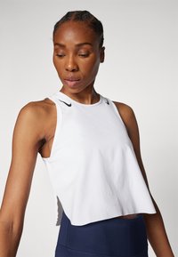 Hvid cropped tanktop med ærmeløst design, har Nike-logo i sort på skuldrene, blødt stof og sideskår for åndbarhed.