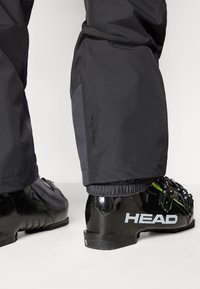 Bottes de ski noires avec la marque "HEAD" portées avec un pantalon étanche noir portant l'étiquette "Helly Tech Protection".