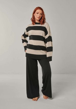 Frau mit roten Haaren, barfuß stehend, trägt einen lockeren schwarz-beigen gestreiften Pullover und weite schwarze Hosen vor einem einfarbigen Hintergrund.