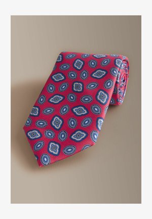 Red silk tie featuring a repeating blue and white paisley pattern, rolled with a smooth texture. Accents include scalloped borders.