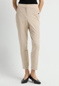 Beige op maat gemaakte broeken met een gladde textuur, voorzien van zijzakken en accenten van gouden knopen. Gecombineerd met zwarte hoge hakken.