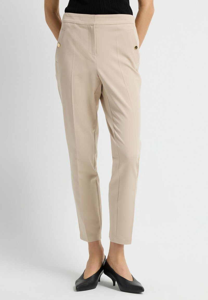 Beige op maat gemaakte broeken met een gladde textuur, voorzien van zijzakken en accenten van gouden knopen. Gecombineerd met zwarte hoge hakken.