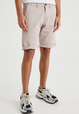 Helle beige Baumwollshorts mit umgeschlagenen Säumen, einem Knopfverschluss und Seitentaschen. Getragen mit grauen und marineblauen Sneakers mit Netz-Details.