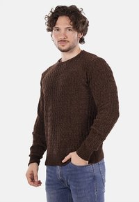 Maglione marrone lavorato a maglia con un motivo testurizzato, scollo rotondo e polsini a costine. Indossato con jeans blu, mostra un look casual.