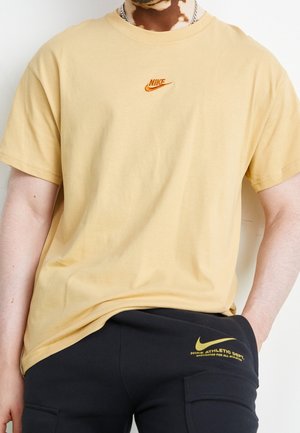 Person iført en beige Nike T-shirt og sorte Nike bukser med synlige logoer, den ene hånd i lommen, foran en lys baggrund.