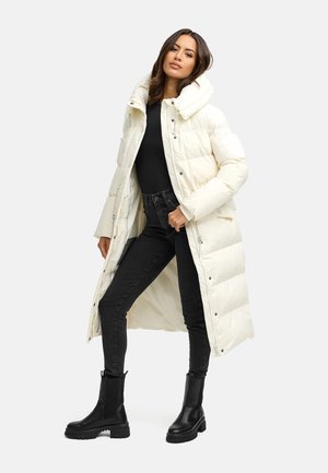 Witte gewatteerde lange jas met hoge kraag, met ritsluiting, gecombineerd met zwarte skinny jeans en zwarte enkellaarsjes, tegen een effen achtergrond.