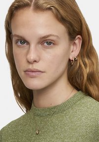 Grøn strikket sweater med en struktureret overflade, lagdelt guldkæde med en dråbeformet vedhæng og hoops øreringe i poleret metal.