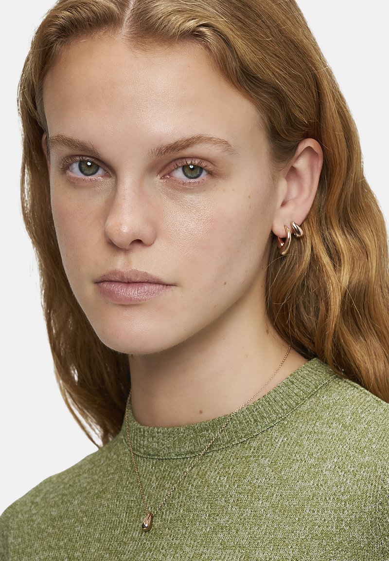 Grøn strikket sweater med en struktureret overflade, lagdelt guldkæde med en dråbeformet vedhæng og hoops øreringe i poleret metal.