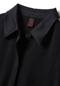 Chemise noire à col avec patte de boutonnage et étiquette rouge et noire indiquant "STEFANEL" cousue à l'intérieur de l'encolure.