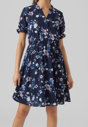 Femme portant une robe fleurie bleu marine avec des manches courtes bouffantes et une taille froncée, debout sur un fond gris uni.