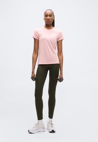 Γυναίκα με ροζ κοντομάνικο μπλουζάκι, σκούρο κολάν και άσπρα αθλητικά παπούτσια, κρατώντας μικρά αλτήρια, στέκεται μπροστά σε απλό λευκό φόντο.