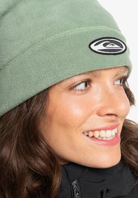 Bonnet en polaire vert avec un logo noir brodé. Texture douce, ajustement confortable et design arrondi, adapté aux activités de plein air.