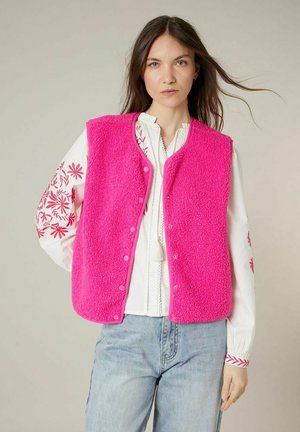 Femme portant une veste sans manches polaire rose vif sur un chemisier blanc avec une broderie florale rouge et un jean bleu clair, debout devant un fond neutre.