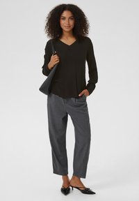 Top nero a maniche lunghe con scollo a V, abbinato a pantaloni grigi sartoriali. La modella tiene una borsa nera e indossa scarpe nere con punta affusolata.