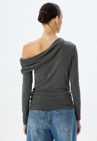 Graues off-shoulder Oberteil mit langen Ärmeln aus drapiertem Stoff; weiche Textur, kombiniert mit hellblauen Jeans, die über Standard-Hintertaschen verfügen.