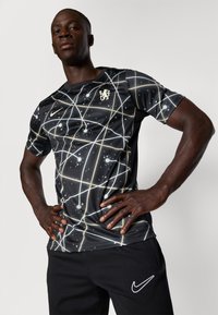 Juoda sportinė marškinė su žvaigždžių raštu balta ir auksine spalvomis, ant krūtinės yra logotipas. Su jais derinamos juodos kelnės ir Nike prekės ženklo akcentas.