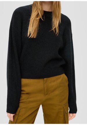 Femme portant un pull noir côtelé à manches longues et un pantalon cargo jaune moutarde, debout devant un fond uni.