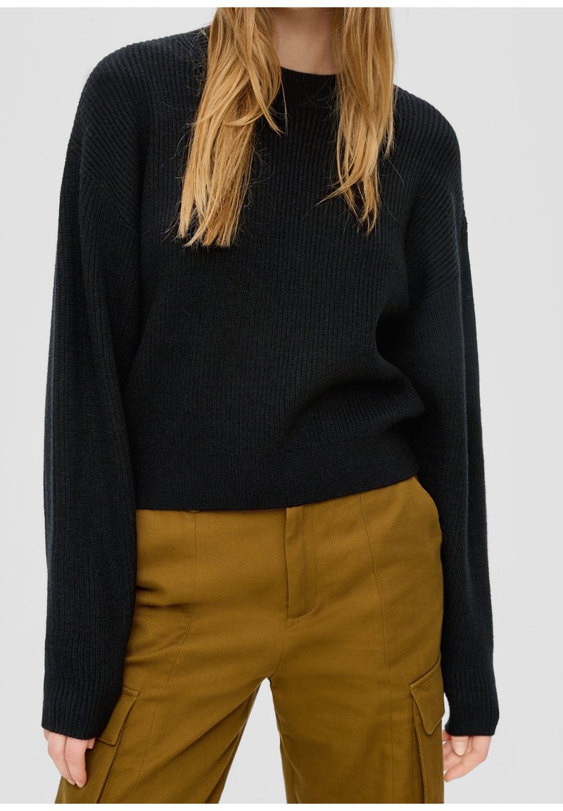 Femme portant un pull noir côtelé à manches longues et un pantalon cargo jaune moutarde, debout devant un fond uni.