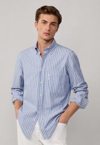Joven con ojos azules que lleva una camisa de botones a rayas verticales en azul claro y blanco, con las mangas arremangadas, con las manos en los bolsillos de unos pantalones blancos.