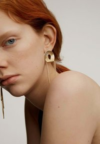 Pendientes circulares de oro con un diseño de espiral, que presentan acentos de cadena alargados, exhibidos en un modelo con cabello rojo y piel clara.