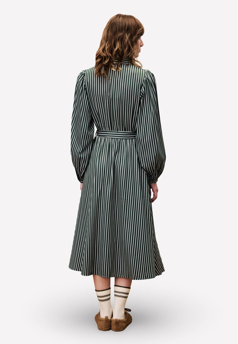 Robe à manches longues rayée verte avec une taille cintrée, une jupe évasée et des rayures verticales. Portée avec des chaussettes rayées à mi-mollet et des pantoufles douces.