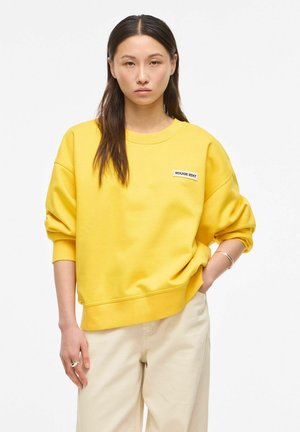 Mlada ženska z dolgimi temnimi lasmi nosi svetlo rumen pulover z napisom "ROUGE EDIT" in svetlo bež hlače, stoji z eno roko v žepu.