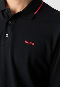 Schwarzes Poloshirt aus Baumwolle mit drei Knöpfen; besitzt einen roten Kragenakzent und ein rotes "HUGO"-Logo auf der linken Brust.