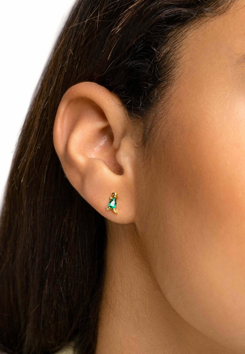 Gros plan sur l'oreille d'une personne portant une petite boucle d'oreille en or avec une pierre précieuse verte en forme de goutte.