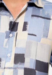 Camicia a maniche corte con bottoni realizzata in tessuto leggero, caratterizzata da un motivo geometrico astratto nei colori blu, beige e nero. I bottoni sono bianchi.