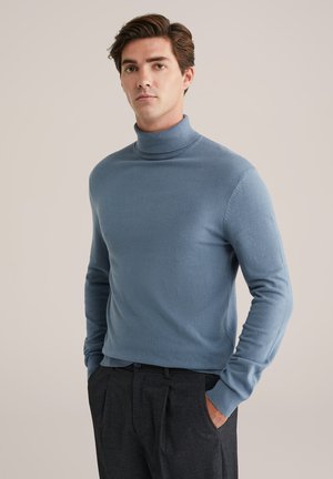 Blauer Rollkragenpullover aus weichem Strickmaterial mit langen Ärmeln, gerippten Bündchen und taillierter Passform. Kombiniert mit dunklen Hosen.