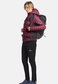 Donna con una giacca a vento bordeaux e pantaloni neri, che indossa uno zaino da trekking nero, in piedi di profilo su uno sfondo bianco.