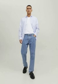 Jovem a caminhar, vestindo uma camisa listrada azul clara sobre uma t-shirt branca, jeans azuis e sapatos pretos, contra um fundo claro e liso.
