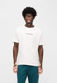 Λευκό βαμβακερό t-shirt με στρογγυλή λαιμόκοψη και κοντά μανίκια, με το κείμενο «New Balance» τυπωμένο σε σκούρο χρώμα στο στήθος. Το μοντέλο φοράει πετρόλ παντελόνια.