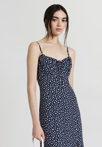 Robe midi bleu marine avec motif floral blanc, fines bretelles, corsage ajusté, devant froncé et fente sur le côté pour une touche supplémentaire.