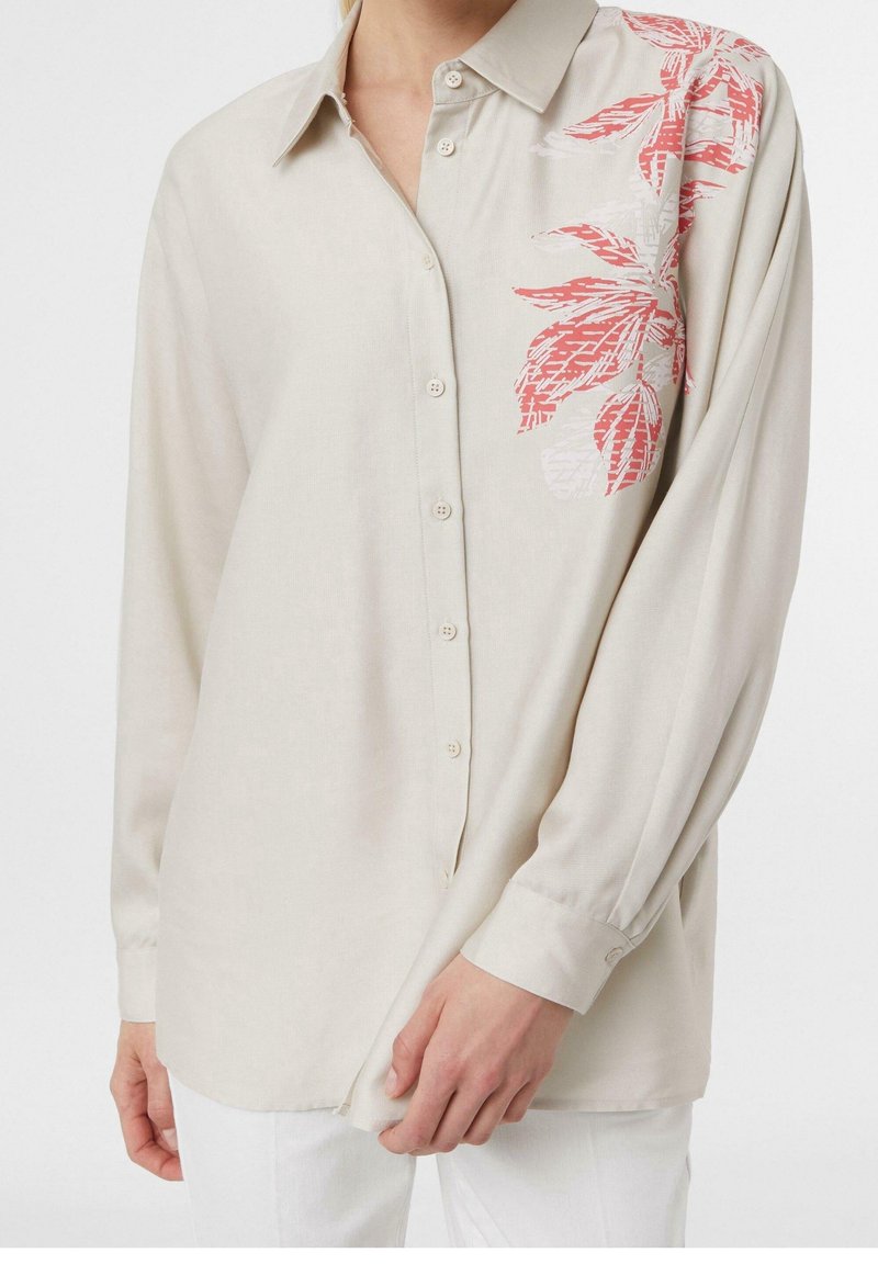 Blouse beige clair à manches longues, boutonnée sur le devant, avec un motif floral rouge sur un côté. Tissu doux et coupe décontractée.