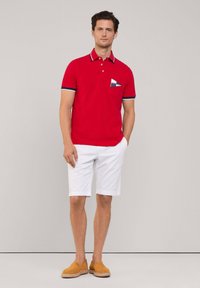 Polo rosso con colletto a righe navy e bianche, con taschino a motivo blu e bianco, abbinato a pantaloncini bianchi e scarpe slip-on color nocciola.
