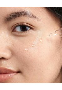 Gouttes de sérum clair sur une surface de peau lisse et claire. La peau semble hydratée avec une lueur naturelle, et une pipette est positionnée près de la joue.