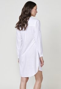 Rochie albă din material ușor, cu mânecă lungă și textură subtilă, având fante laterale, yoke posterior și detalii cu nasturi la manșete.