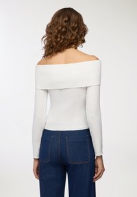 Biała, prążkowana bluzka na ramiączkach z długimi rękawami, zestawiona z ciemnoniebieskimi dżinsami z dwiema tylnymi kieszeniami i pomarańczowymi detalami w szwach.