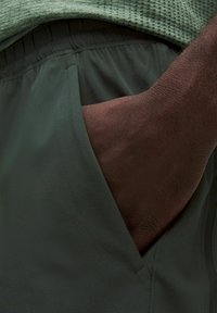 Mörkgröna sportshorts med en slät yta, utrustade med en sidoficka. En hand är placerad i fickan, vilket visar hudton.