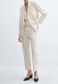 Traje beige a medida con blazer de doble botonadura, pantalones ajustados y chaleco a juego. Acentuado con mules negros de punta.
