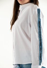 Camisa blanca con cuello mao, que presenta un acento desgastado en azul claro a lo largo de la mangas con letras negras que dicen "QUIÉRETE A TI MISMO".