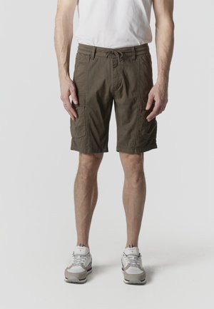 Mężczyzna ubrany w brązowe cargo shorts z wieloma kieszeniami, białe trampki i białą koszulkę na jasnym, jednolitym tle.