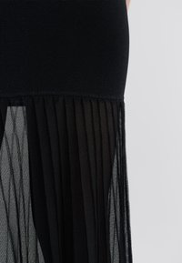 Rochie neagră cu un top strâmt din material texturat, care se transformă într-o fustă plisată dintr-un material transparent, stratificată, la baza rochiei.