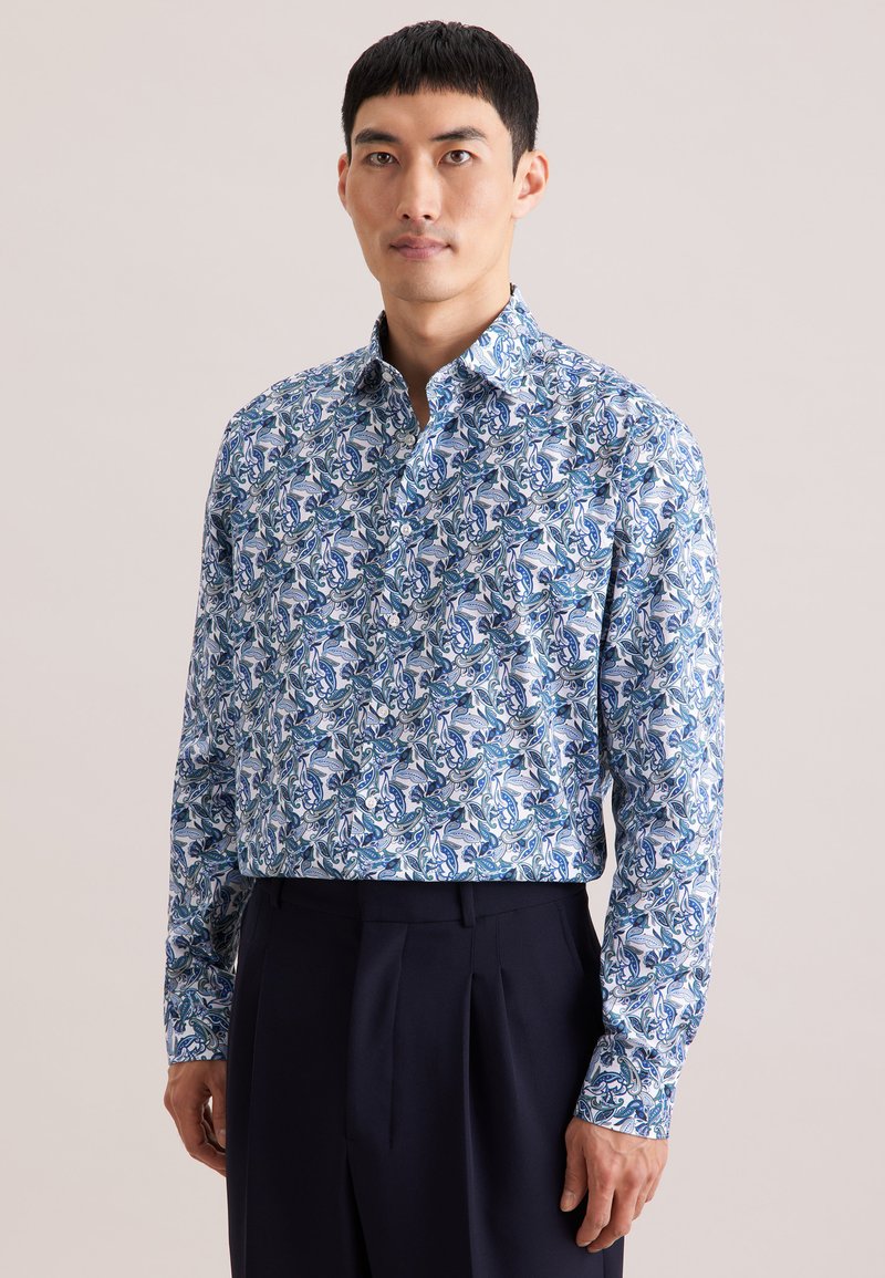 Langarmshirt met een blauw-wit bloemenpatroon op lichtgewicht stof. Klassieke kraag en knoopsluiting aan de voorkant, gecombineerd met een donkere broek.